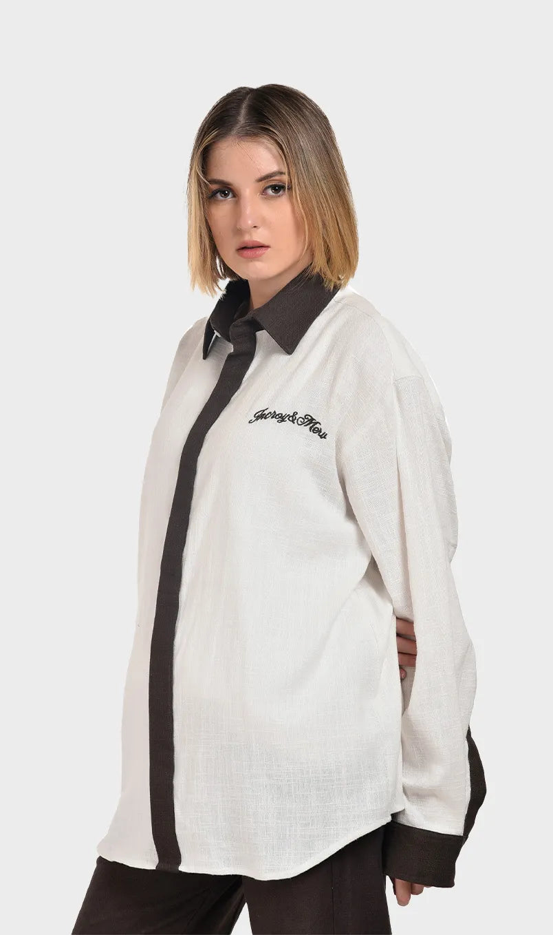 Three-quarter front view of a female model wearing an oversized off-white slub cotton button-down shirt with brown trim.