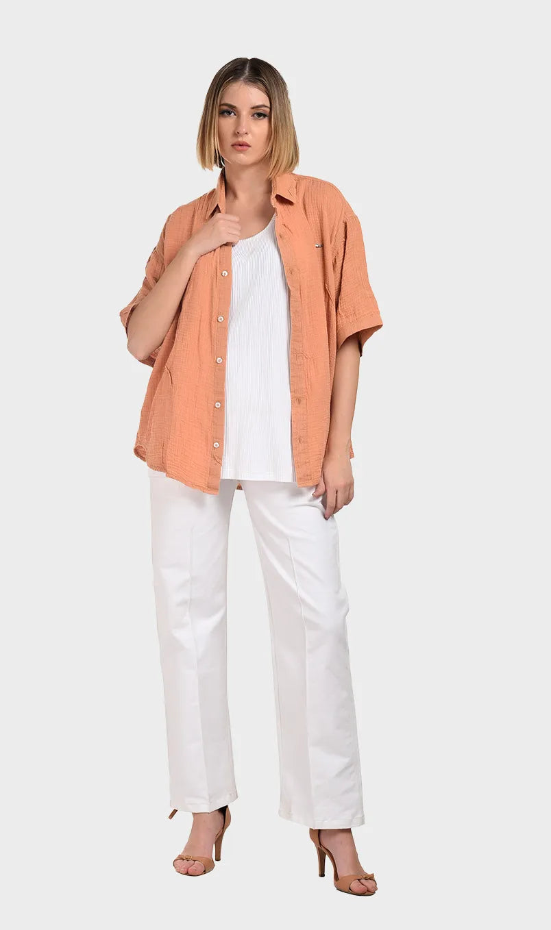 Full-body shot of a model wearing an open peach 100% cotton double cloth short-sleeve shirt over a white tank top and white trousers.
