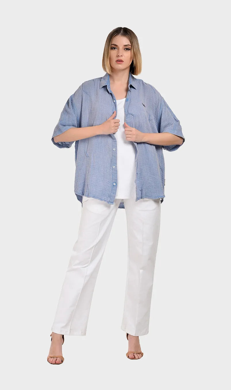 Full-body shot of a model wearing an unbuttoned light blue 100% cotton double cloth shirt layered over a white top with white straight-leg trousers.