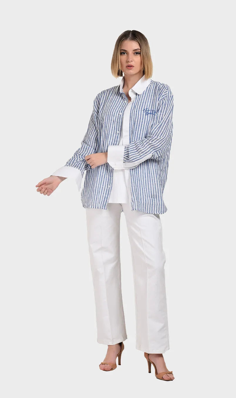 Full-body view of a model in an unbuttoned blue striped dobby fabric shirt paired with white wide-leg trousers and heels.