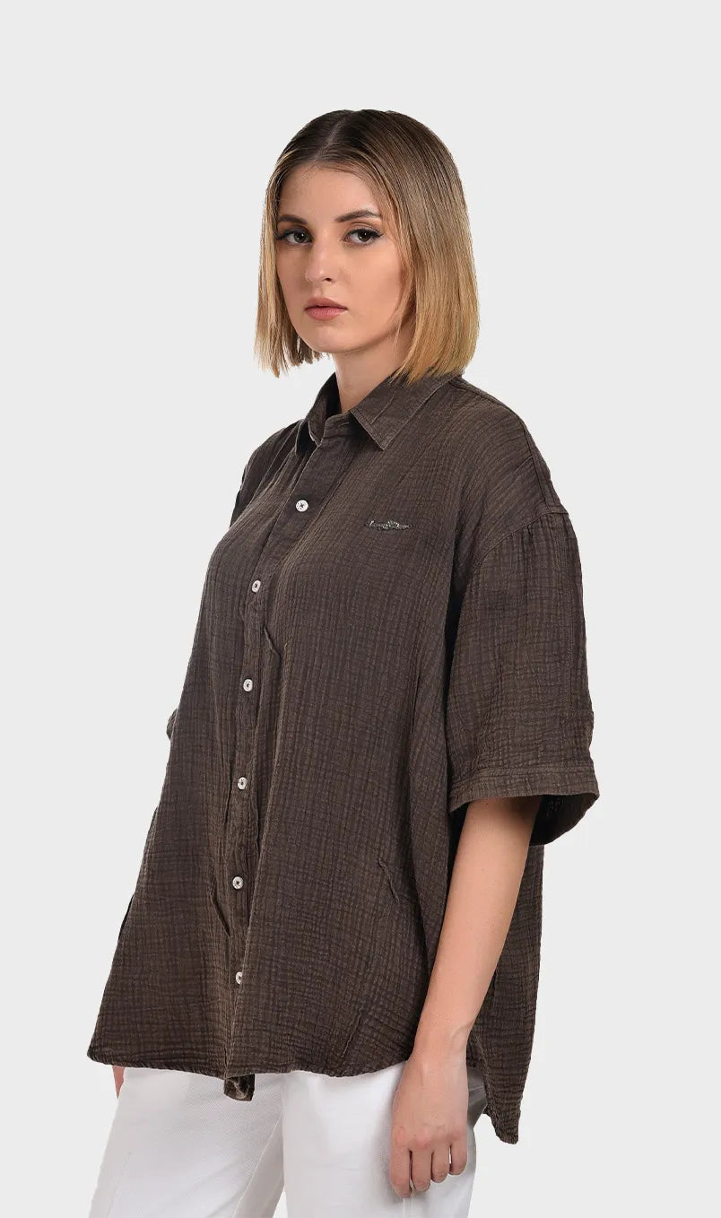 Three-quarter left angle showing the chest embroidery detail and relaxed drop sleeves on a women's brown cotton double cloth top.