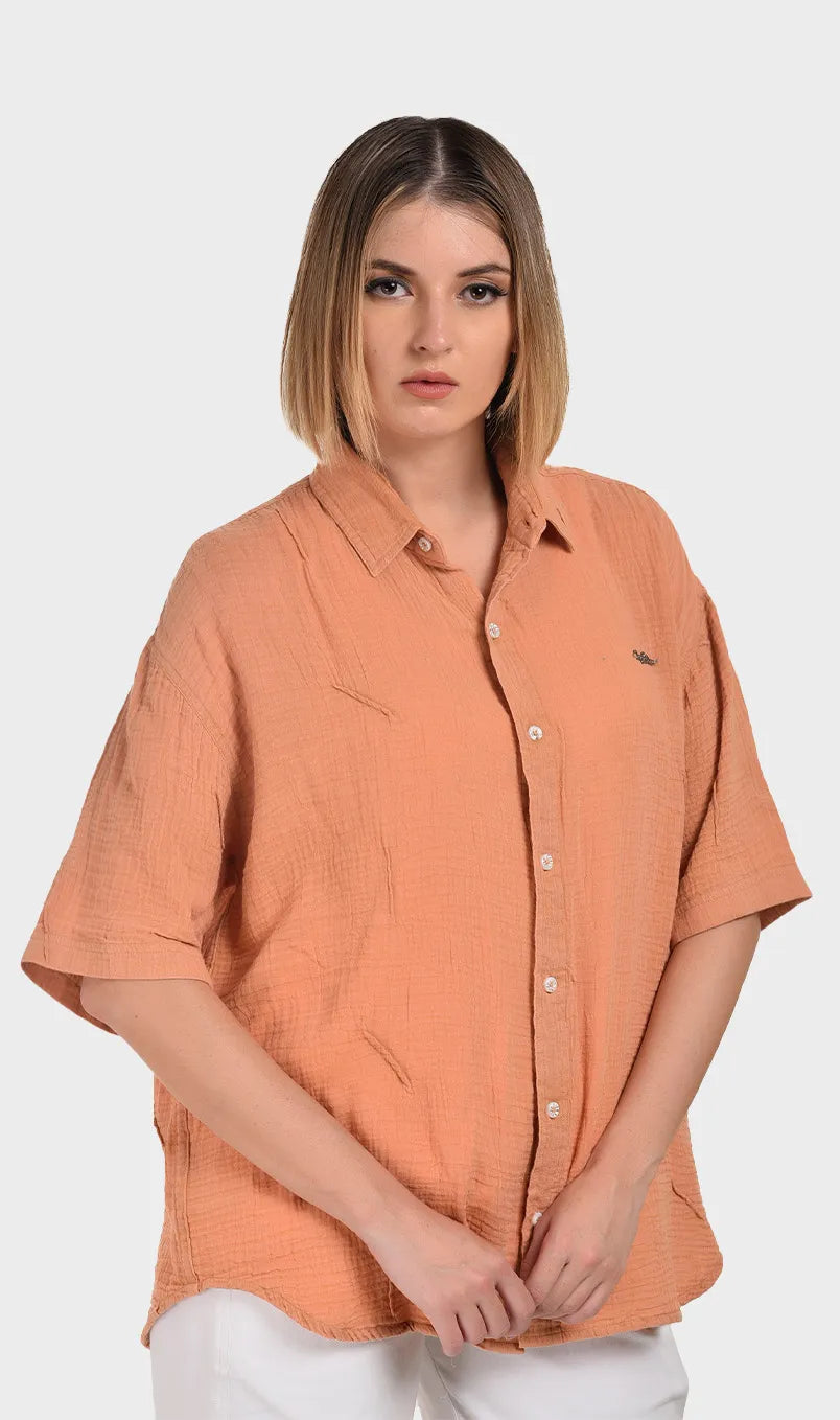 Half-turn front view highlighting the chest logo detail and drop-shoulder sleeves on a women's peach 100% cotton double cloth shirt.